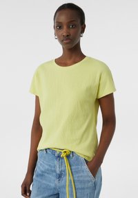 Koszulka z krótkim rękawem w jasnym żółtym kolorze o teksturowanym wyglądzie. Połączona z jasnoniebieskimi dżinsami i zawiązanym żółtym paskiem.