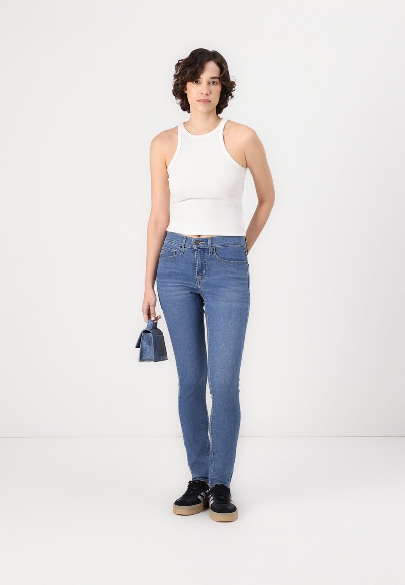 Débardeur blanc ajusté en côte, jeans skinny bleus, baskets noires avec semelles beige, tenant un petit sac bleu. Design minimaliste, style décontracté.