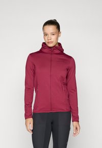 Frau, die einen engen weinroten Reißverschluss-Hoodie trägt, mit entspannten Händen an den Seiten, vor einem schlichten hellen Hintergrund.