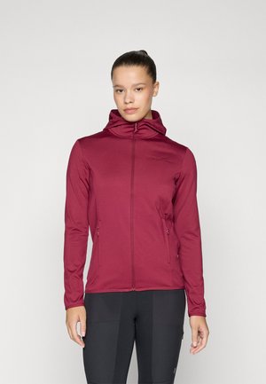 Vrouw in een aansluitende bordeauxrode zip-up hoodie met ontspannen handen naast haar lichaam, tegen een effen lichte achtergrond.
