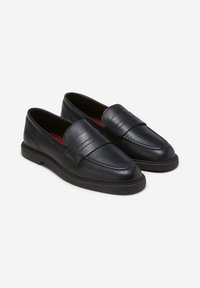 Sorte læder slip-on loafers med afrundede tæer, en stropdetalje på toppen, og sorte gummisulder mod en ensfarvet hvid baggrund.