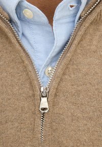 Beige gebreide trui met een gedeeltelijke rits, gedragen over een lichtblauw overhemd met kraag. Heeft subtiele textuur en zilveren ritsbeslag.