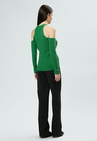 Grünes geripptes Langarmoberteil mit Cold-Shoulder-Design, kombiniert mit schwarzen weiten Hosen; glatte Textur, figurbetonter Schnitt.