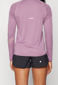 Nainen, joka on pukeutunut mauven väriseen pitkähihaiseen urheilu-paitoihin, joissa on heijastavat yksityiskohdat, ja mustiin juoksushortseihin, seisoo selin.