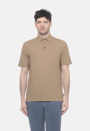 Uomo con capelli corti e scuri, indossa una polo a maniche corte color marrone chiaro e pantaloni blu, in piedi dritto contro uno sfondo chiaro e uniforme.