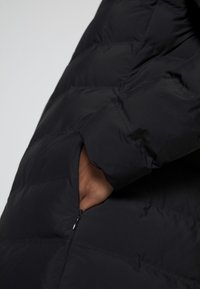 Chaqueta acolchada negra con textura suave, bolsillo con cremallera y diseño acolchado, que destaca las propiedades de calor e aislamiento.