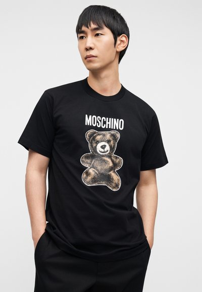 Μαύρο μπλουζάκι με στάμπα αρκούδας σε καφέ και λευκό χρώμα, με το κείμενο "MOSCHINO" πάνω από το σχέδιο. Κοντά μανίκια και λαιμόκοψη στρογγυλή.