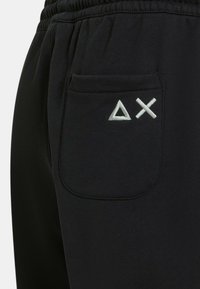 Pantaloni della tuta neri con una fascia elastica in vita e una tasca posteriore con dettagli ricamati bianchi a triangolo e croce. Tessuto dalla texture liscia.