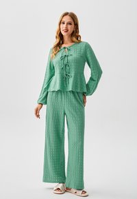 Completo verde strutturato in due pezzi con top peplum a maniche lunghe e dettaglio con nodo, abbinato a pantaloni a gamba larga e sandali open-toe.