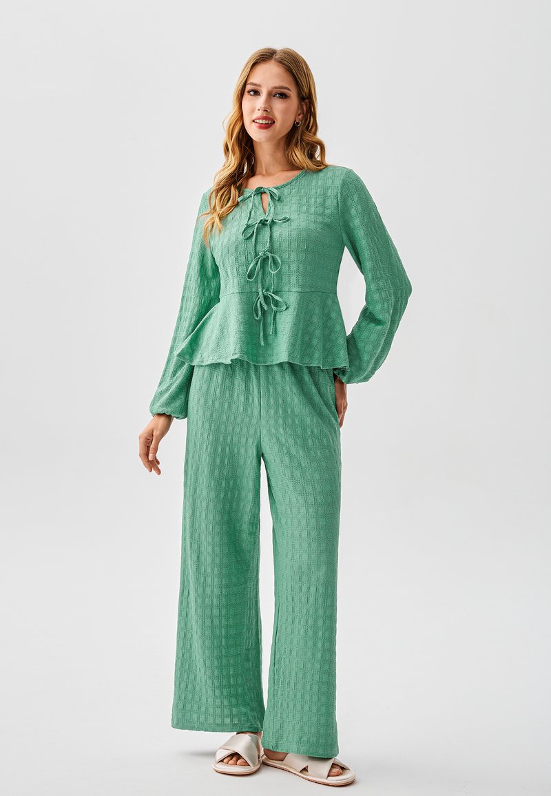 Completo verde strutturato in due pezzi con top peplum a maniche lunghe e dettaglio con nodo, abbinato a pantaloni a gamba larga e sandali open-toe.