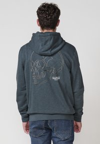 Sötét szürke kapucnis pulóver, puha anyagból, fehér keretes koponya mintával a hátsó részén. Húzózsinóros kapucnija és bordázott mandzsettái és alja van.