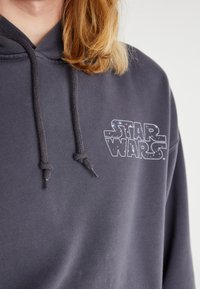 Sötét szürke kapucnis pulóver, állítható fűzővel, fehér és kék galaxismintás "Star Wars" logóval a mellkasán. Puha szövettextúra.