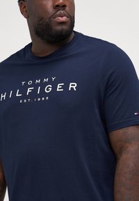 Tumši zila kokvilnas T-krekls ar "TOMMY HILFIGER" logo baltā krāsā. Ar apaļu kakla izgriezumu, īsām piedurknēm un mazu karoga akcentu uz piedurknes.