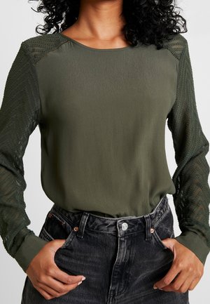 Kvinde iført olivengrøn bluse med lange ærmer og teksturerede, gennemsigtige ærmer samt sorte højtaljede jeans, hænderne i lommerne.