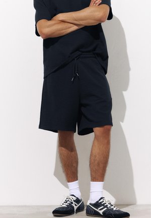 Personne portant un short noir et des baskets noires avec des lacets blancs et de hautes chaussettes blanches, debout, les bras croisés, contre un mur blanc.