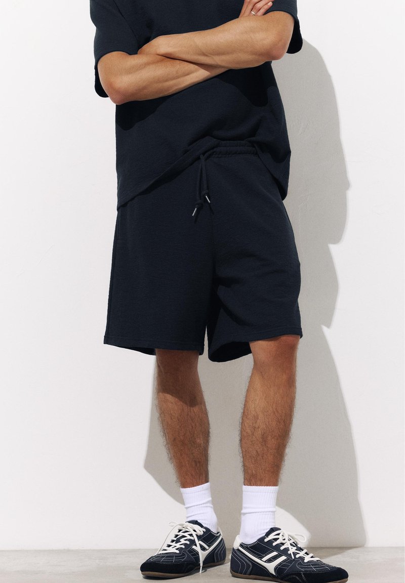 Personne portant un short noir et des baskets noires avec des lacets blancs et de hautes chaussettes blanches, debout, les bras croisés, contre un mur blanc.