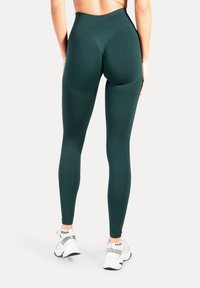 Leggings a vita alta di colore verde scuro con una texture liscia e una vestibilità aderente, abbinati a scarpe sportive bianche con accenti riflettenti.