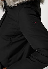 Manche de veste d'hiver noire avec écusson logo Tommy Hilfiger, capuche bordée de fourrure et bouton-pression argenté près du col.