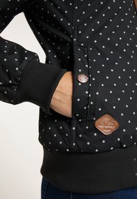 Černá bomberka s bílými puntíky, hladk�ými žebrovanými manžetami a koženou nášivkou s logem. Obsahuje boční kapsu se zavíráním na patent.