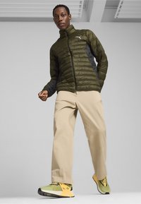 Puffjakke i olivengrøn med sorte sider, beige bukser og multicolor sneakers med gule og grønne accenter.