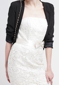 Svart kavaj med kedjesnor, över en vit spets klänning utan axelband med ett satinband i midjan, som visar blomstertryck och figurnära silhuett.