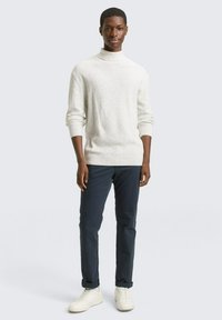 Un pull à col roulé blanc à motifs, ajusté, porté avec un pantalon bleu foncé et des baskets blanches, présenté sur un modèle sur fond clair.