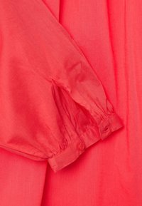Blusa rossa di cotone con maniche lunghe, polsini arricciati e dettaglio con bottone al polso. Texture liscia e design morbido.