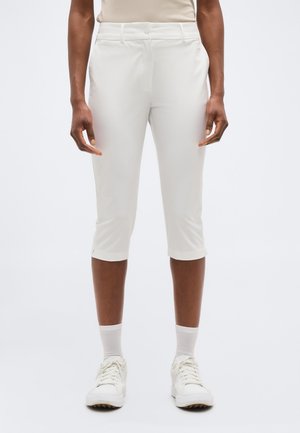 Άτομο που φοράει λευκό παντελόνι λίγο πάνω από τον αστράγαλο, λευκές κάλτσες μέχρι τον αστράγαλο και λευκά αθλητικά παπούτσια, στέκεται με τα χέρια χαλαρά στα πλάγια.