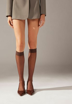 Brune knæhøje strømper med en gennemsigtig tekstur, prydet med en solid brun kant øverst, som bæres med spidse brune hæle og en grå blazer.