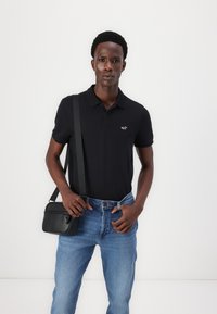 Ung mann iført svart polo-skjorte, lyseblå jeans og svart skulderveske foran en ensfarget hvit bakgrunn.