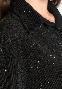 Black sequined jacket featuring a ribbed texture, button details, and a structured collar. The fabric has a shimmering effect.
