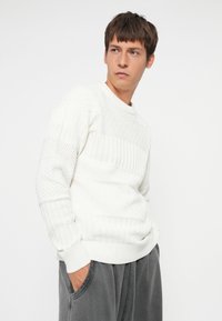Hvid strikket trøje med teksturerede mønstre, ribbede manchetter og udskæring. Modellen bærer grå bukser med sidelommer. Enkel og moderne design.