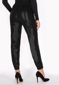 Zwarte faux leren joggers met een glanzende afwerking, een elastische tailleband, zijzakken en taps toelopende boorden, gedragen met zwarte hoge hakken.