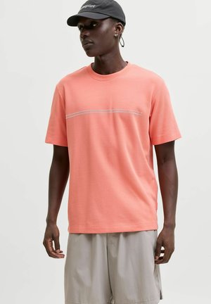 Homme portant un t-shirt corail à manches courtes avec une fine rayure horizontale et un short gris clair, casquette noire, debout devant un fond uni.