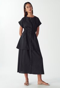 Vestito midi nero con maniche corte, apertura frontale con bottoni, vita elastica, design a strati e tessuto liscio. Indossato con semplici sandali piatti.