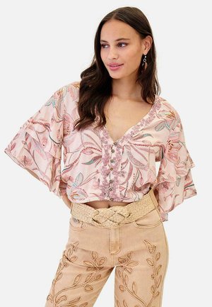 Mujer con blusa rosa de flores con mangas con volantes y pantalones beige con patrones tejidos de hojas, posando con las manos en las caderas.