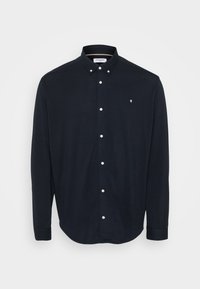 T-paita tummansinisestä pehmeästä kankaasta, jossa on kaulus, pitkät hihat ja valkoiset napit edessä. Pieni logo rinnassa.
