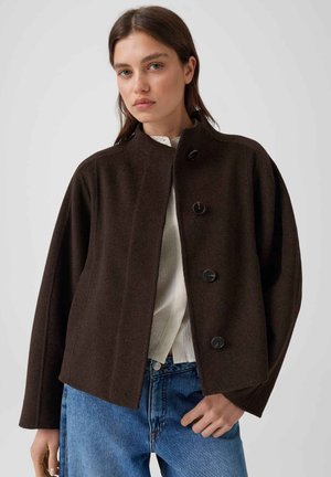 Donna che indossa un cappotto marrone scuro abbottonato sopra una camicia bianca e jeans blu su uno sfondo chiaro uniforme.