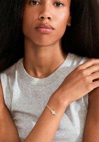 Zilveren armband met bloemenmotief, dunne ketting, gedragen aan een pols. Achtergrond toont een lichtgrijs mouwloos topje. Glad huidoppervlak.