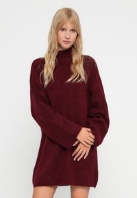 Bordo prevelik pulover z visokim ovratnikom, rebrasto teksturo in širokimi rokavi, izdelan iz mehkega pletenega materiala.
