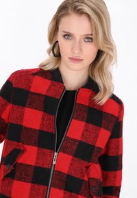 Bomber jacket a quadri rossi e neri con chiusura a zip, tasche laterali e colletto a coste. Realizzato in un tessuto a maglia testurizzato.