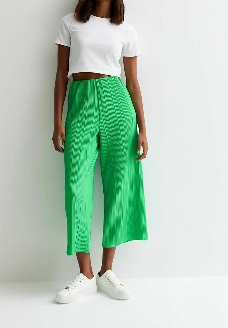 Woman wearing a white cropped t-shirt, bright green wide-leg pleated pants, and white sneakers, standing against a plain white background.