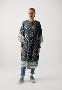 Dlhý, vzorovaný kimono s geometrickými motívmi v čiernej a bielej farbe. Má široké rukávy, viazanie v páse a viacvrstvové textúry. Nosené cez modré džínsy.