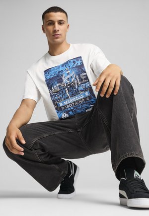 Hvid t-shirt med et blåt grafisk print med tekst og et logo. Mørke jeans og sorte sneakers fuldender outfittet. Glat, afslappet pasform.