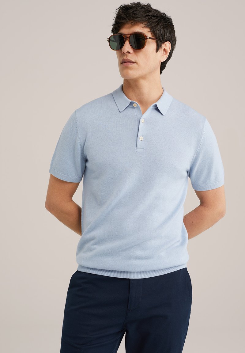 Cămașă polo de culoare albastru deschis, cu mâneci scurte și guler, având un sistem de închidere cu trei butoni și textură netedă. Pur și simplu asortată cu pantaloni negri și ochelari de soare.