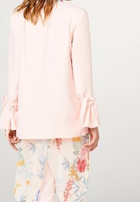 Kobieta ubrana w jasnoróżową marynarkę z związanymi detalami rękawów, w pastelowej spódnicy w kwiatowy wzór, pokazana z tyłu.