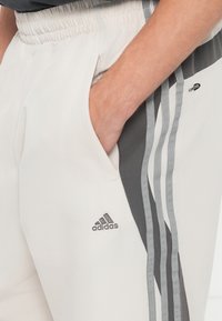Adidas sporta bikses gaiši krēmkrāsā ar pelēkiem sānu svītrām, ar elastīgu jostu un mazu logotipu gurnā. Gluda tekstūra.