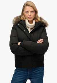 Sort pufferjakke med pelsforet hætte, frontknapper og ribbede manchetter; båret over en lysefarvet sweater og blå jeans.