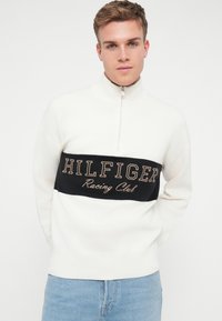 Balts džemperis ar augstu apkakli, rāvējslēdzēju un melnu svītru ar zelta izšuvumu "HILFIGER Racing Club". Mīksta tekstūra.