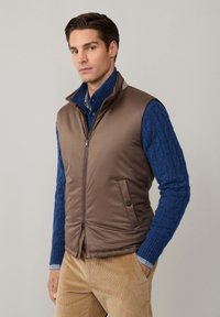 Gilet imbottito marrone con colletto alto, pannelli cuciti e tasche laterali sopra un maglione blu a trecce, abbinato a pantaloni in velluto a coste color cammello.